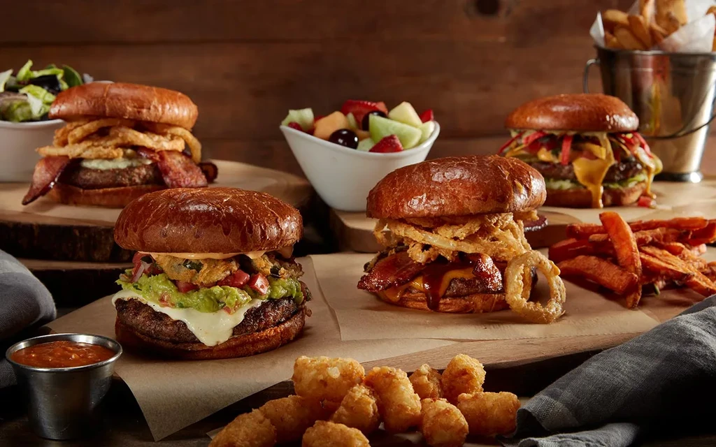 Four gourmet burgers, fries, tater tots, salads, and fruit on wood.