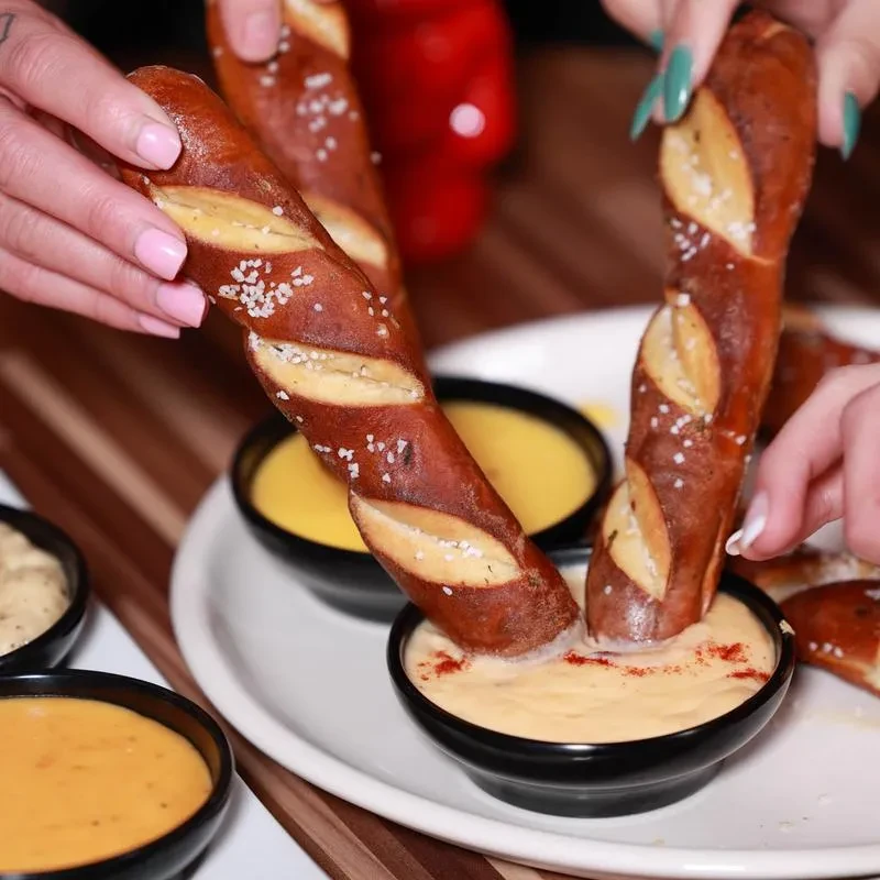 Two hands dipping salted soft pretzels into bowls of cheese sauce.
