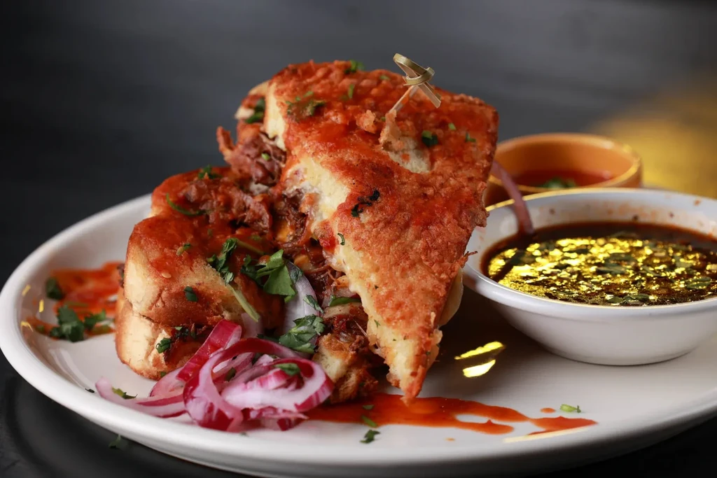 Hearty toasted sandwich with red sauce, green herbs, and two dipping bowls.