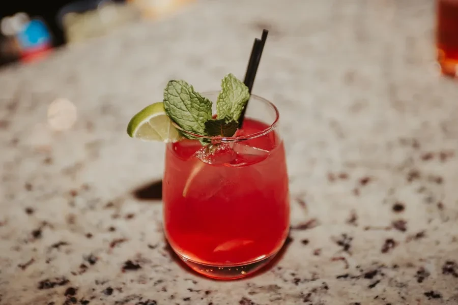 Red cocktail in a clear glass with lime, mint, ice, and straws.