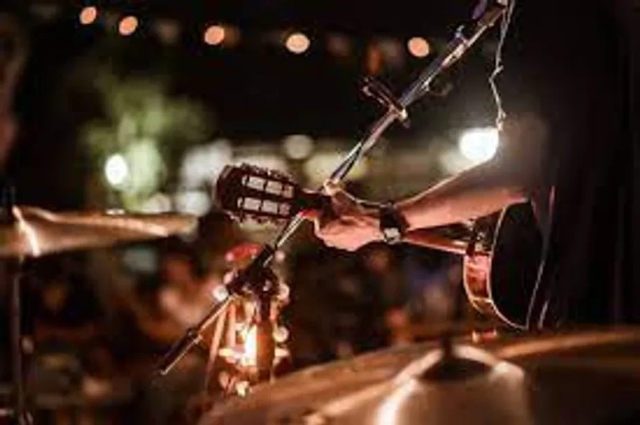 Musician's hand strumming acoustic guitar onstage with microphone and cymbals.