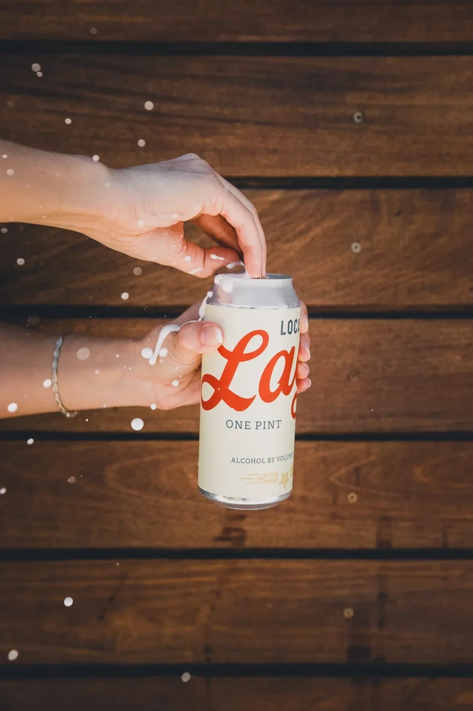 Hands opening 'LOCAL LAGER' can, white liquid splashing and dripping.