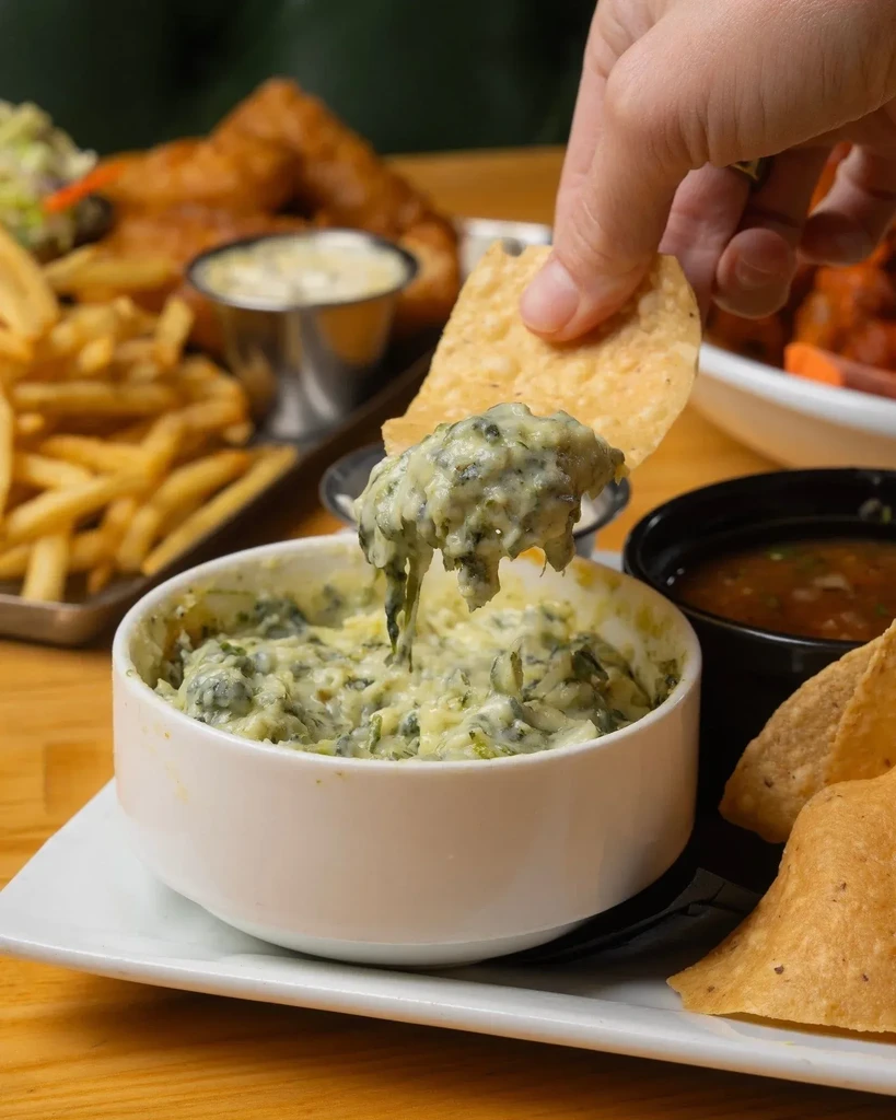 Hand dips tortilla chip into gooey spinach artichoke dip on wooden table.