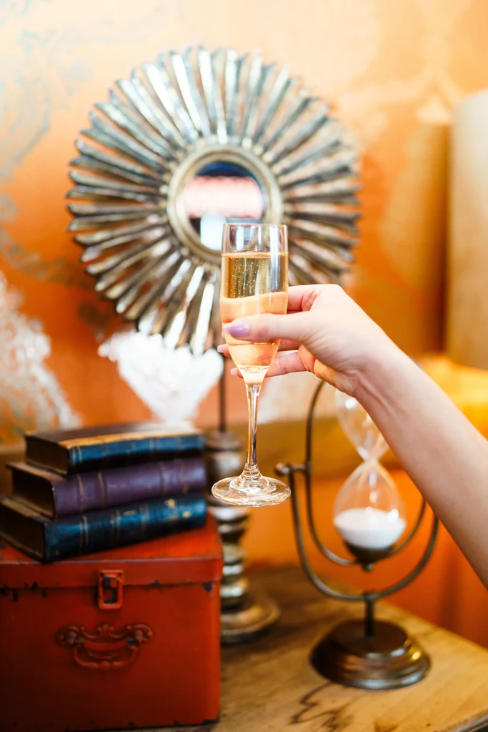 Manicured hand with purple nails holds sparkling wine flute in elegant interior.