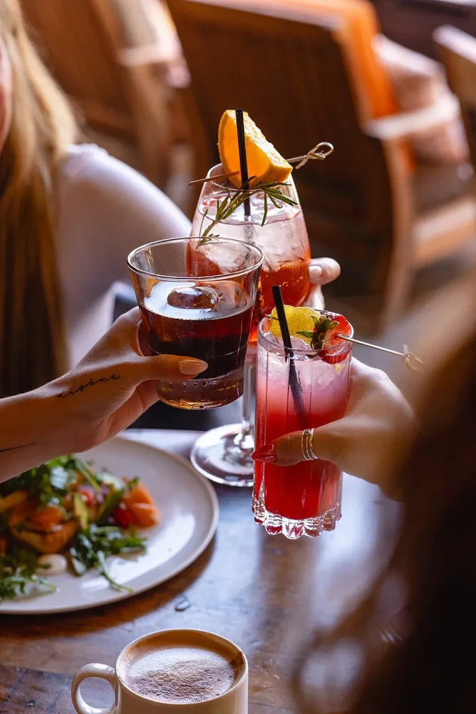 Three hands toast vibrant cocktails and coffee at a restaurant table.