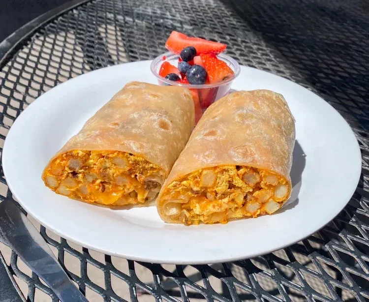 Cut breakfast burrito with eggs, potatoes, cheese, and fruit cup outdoors.
