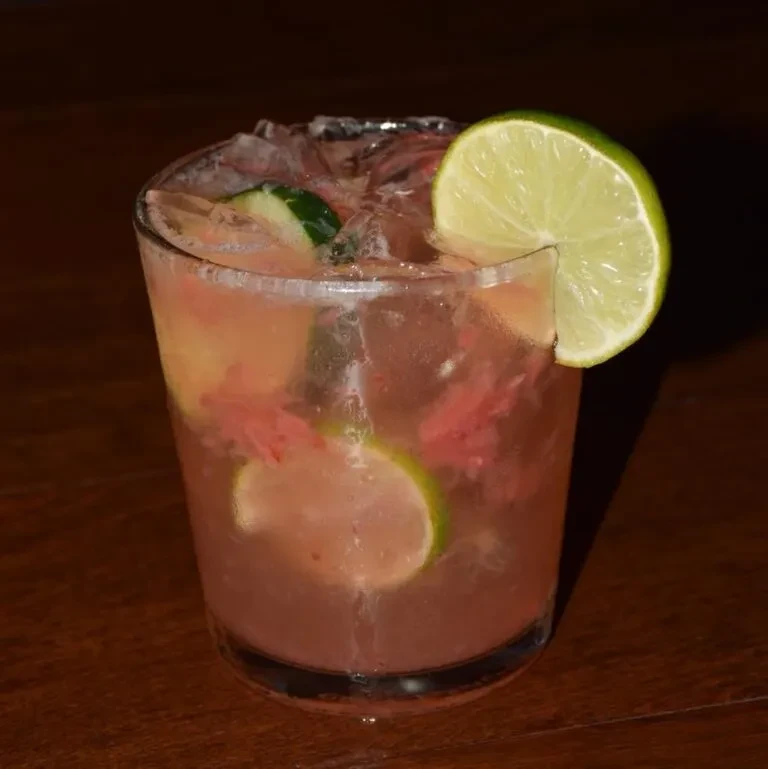 Reddish-pink drink with ice, cucumber, lime, and flower petals on wood.