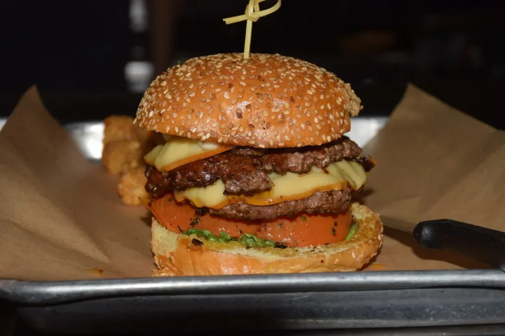 Double-patty burger with cheese, tomato, greens, and skewer on a tray.
