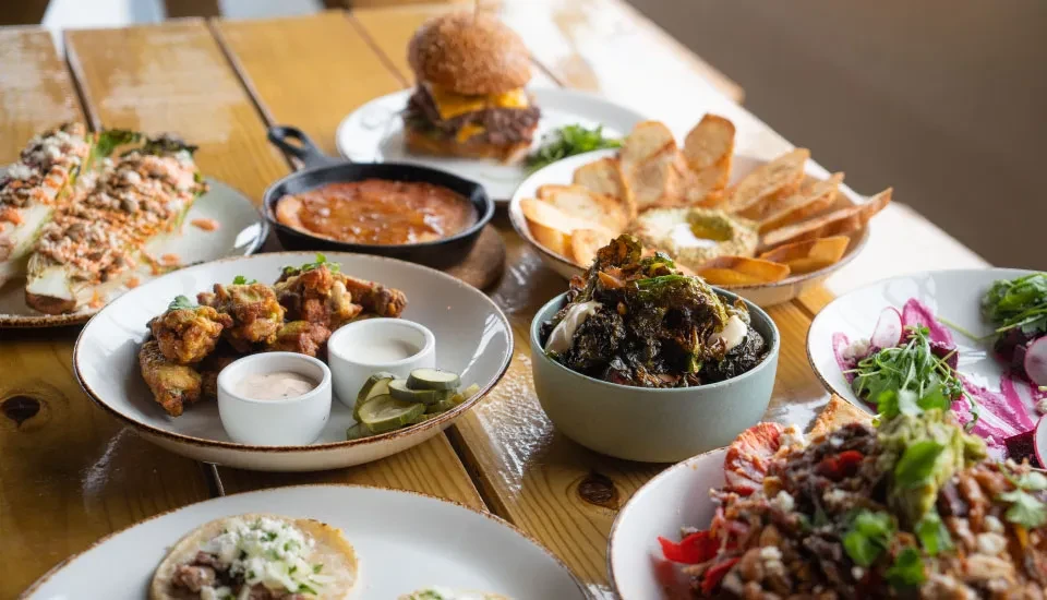 Table with diverse plates: tacos, wings, cheeseburger, and salads.