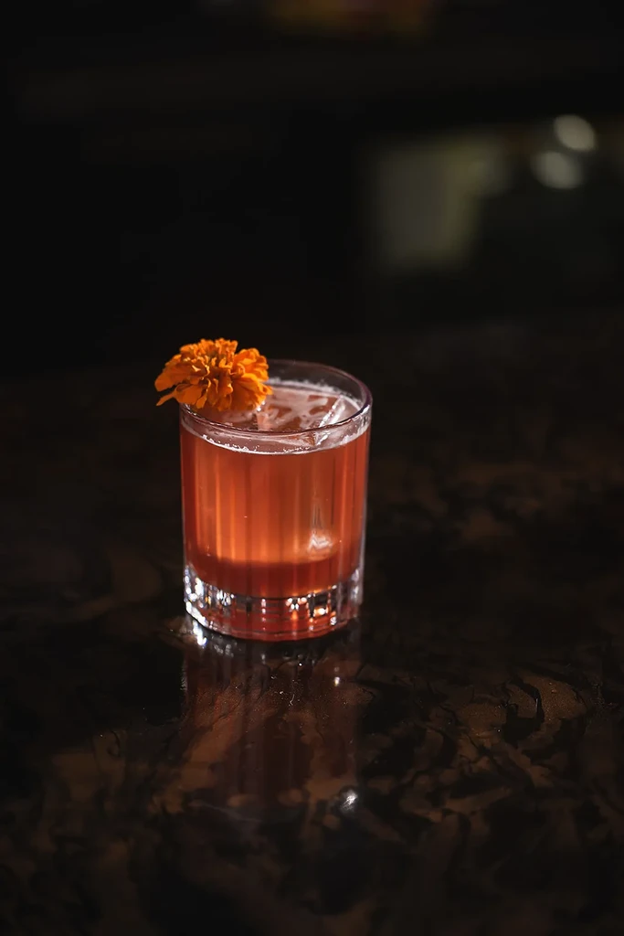 Red-orange cocktail in ribbed glass, garnished with an orange marigold flower.