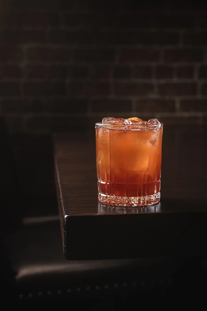Reddish-brown cocktail with ice, citrus peel, in rocks glass on wood.