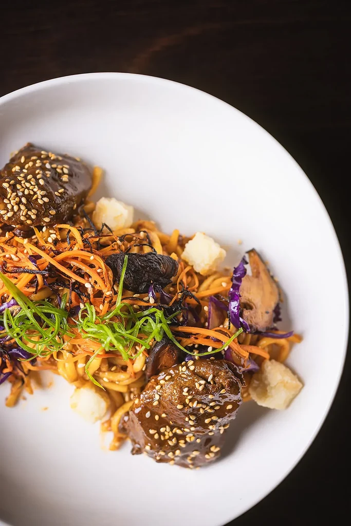 Plated Asian noodles with meat, colorful vegetables, sesame seeds, and dark sauce.