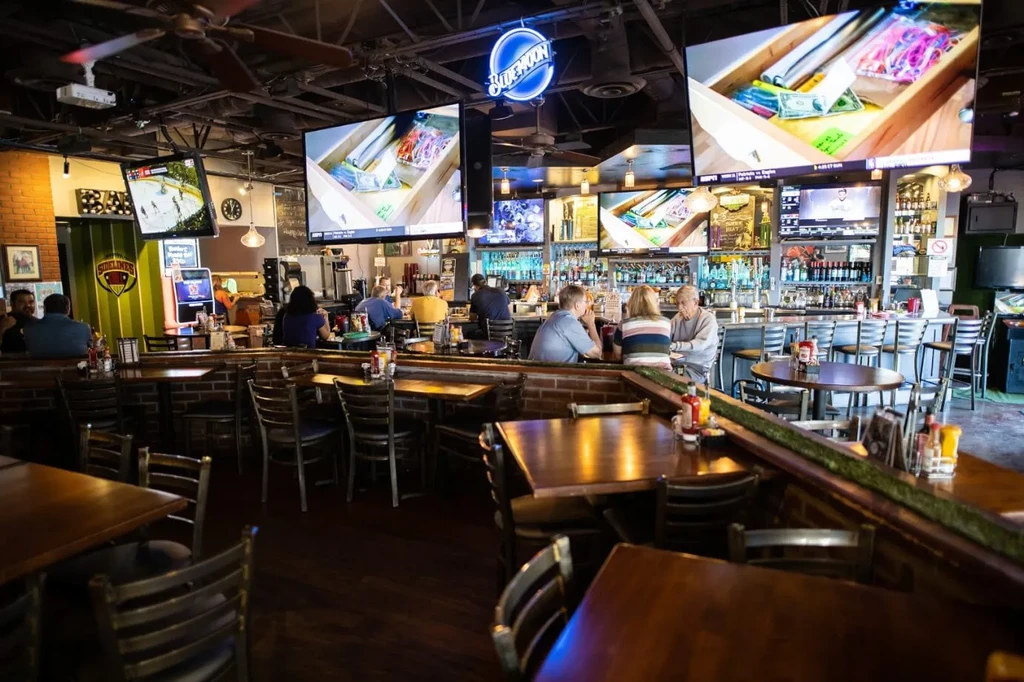 Lively sports bar interior with wooden tables, a long bar, and TVs.