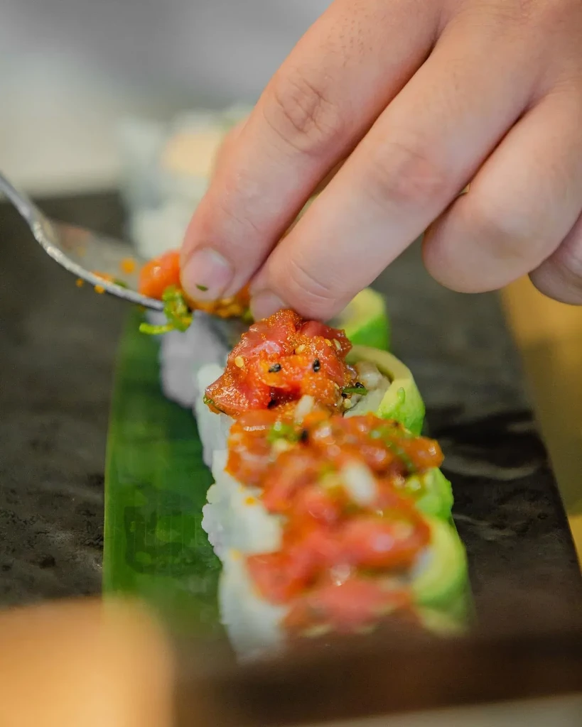 Chef's hand garnishes sushi rolls with red tuna poke on a dark platter.