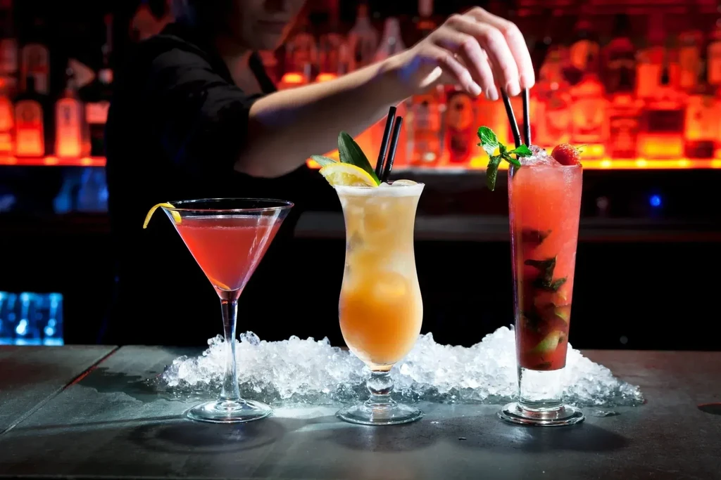 Garnished cocktails on a bar with crushed ice; bartender adjusts a drink.