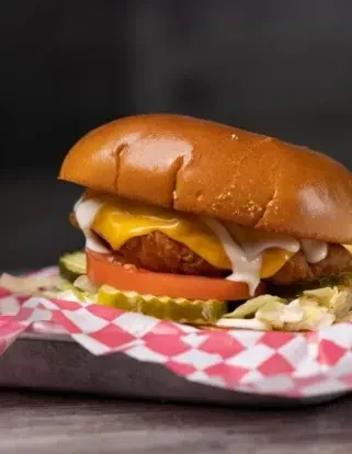 Bun sandwich with melted cheese, tomato, pickles, lettuce on checkered paper.