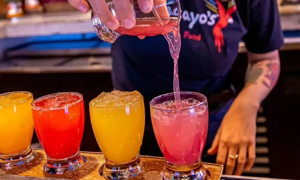 Hand pouring pink drink among four colorful beverages on counter.