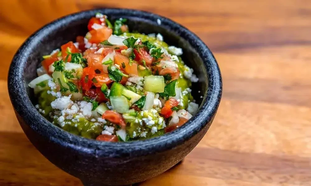 Rustic bowl of guacamole topped with tomatoes, onions, cilantro, crumbled cheese.
