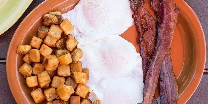 Breakfast meal with three fried eggs, cubed potatoes, and crispy bacon.