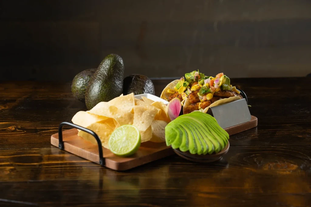 Two colorful tacos, chips, lime, and avocado on a dark wooden table.