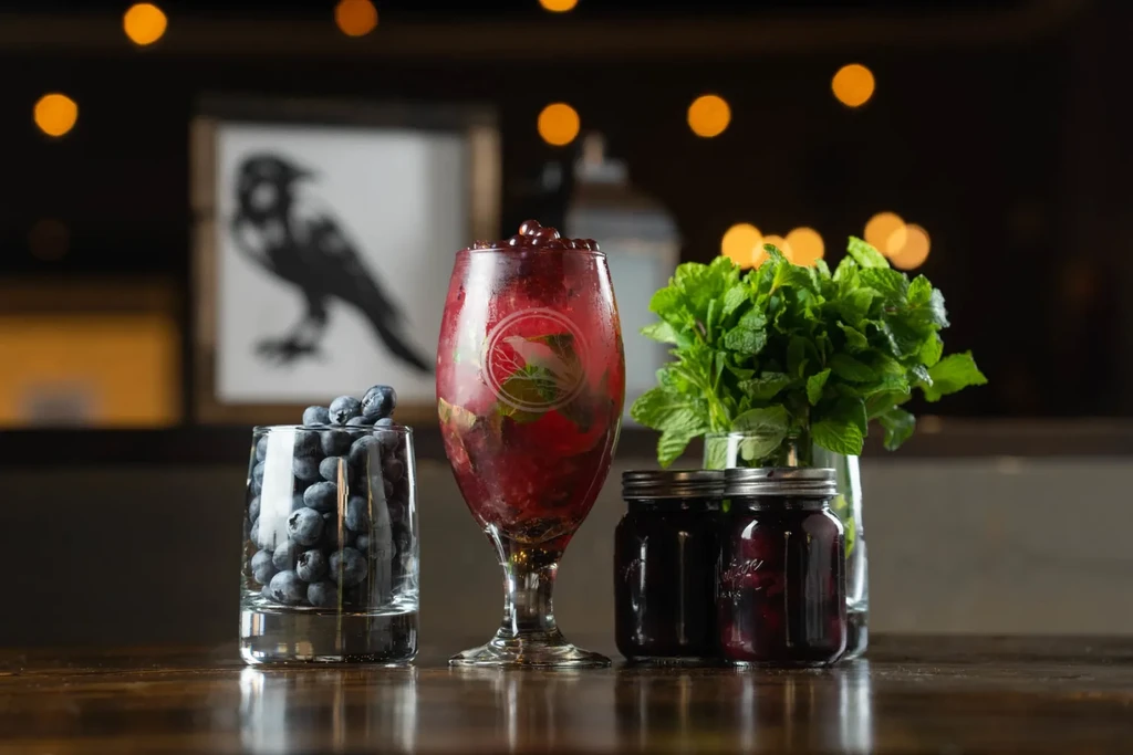 Still life of red berry drink with blueberries, mint, and preserves on wood.