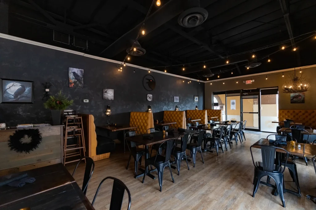 Empty restaurant with dark tables, mustard booths, raven art, and warm lights.