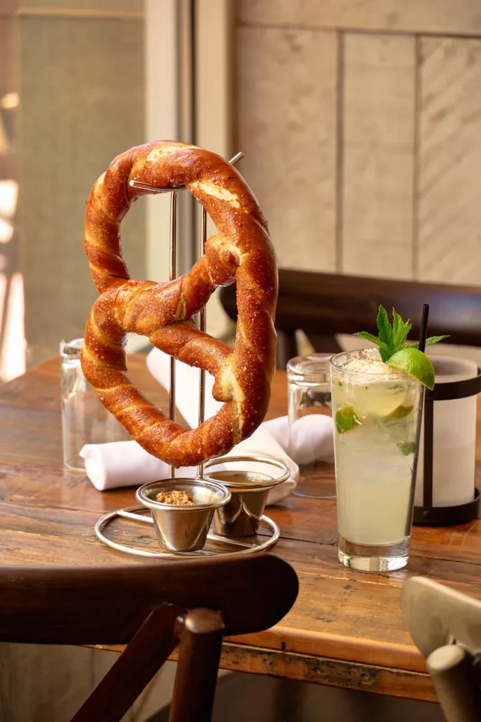 Fresh pretzel, mustard, dip, and a lime cocktail on a wooden table.