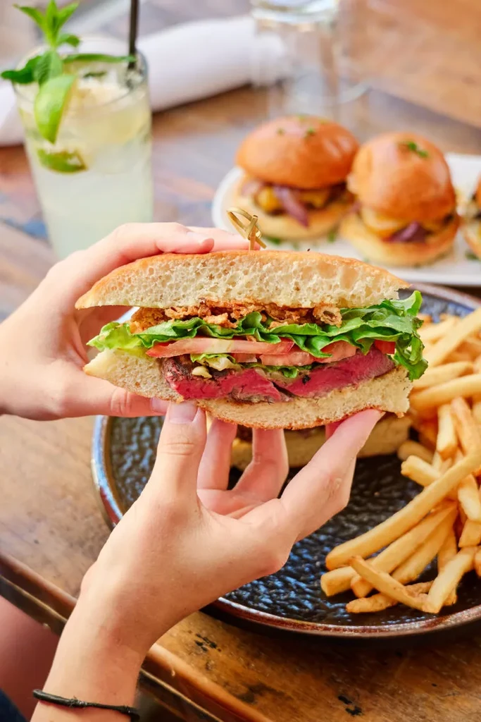 Hands holding a sliced steak sandwich with fries and mini-burgers.