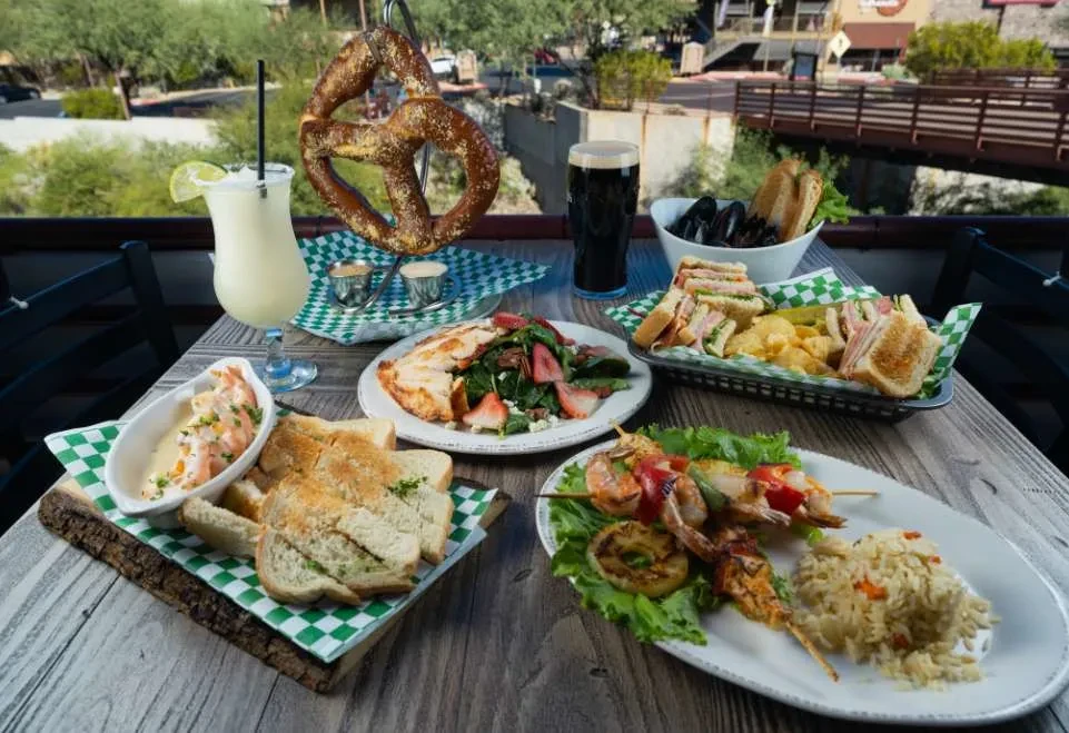 Outdoor dining table with diverse food, drinks, and a scenic background.