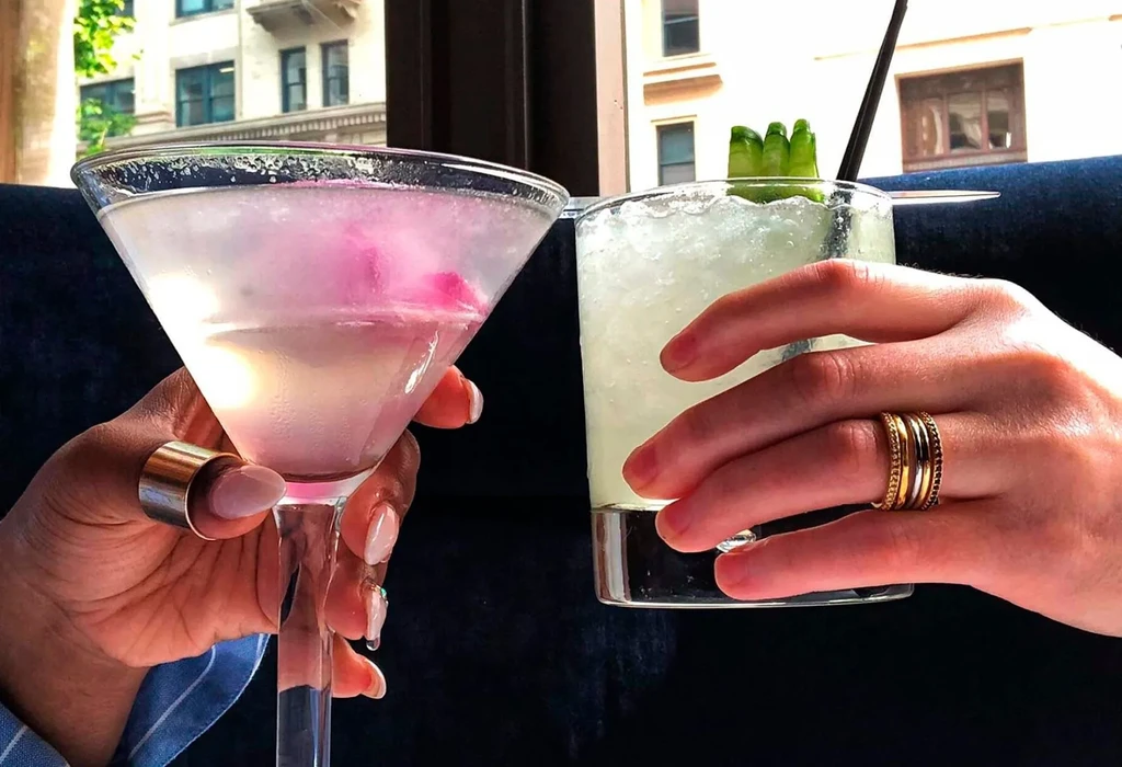 Two hands holding a pink martini and a green cucumber cocktail.