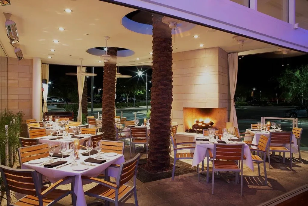 Empty upscale outdoor patio with set tables, warm lighting, lit fireplace.