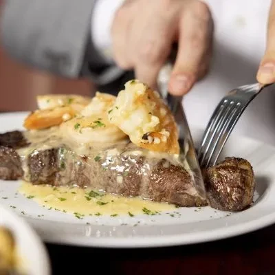 Hands cutting gourmet steak and shrimp with creamy sauce.