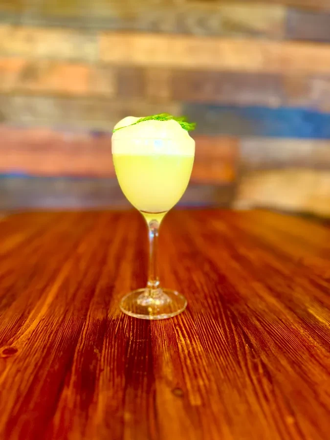 Yellow-green cocktail with foam and herb on wooden table.