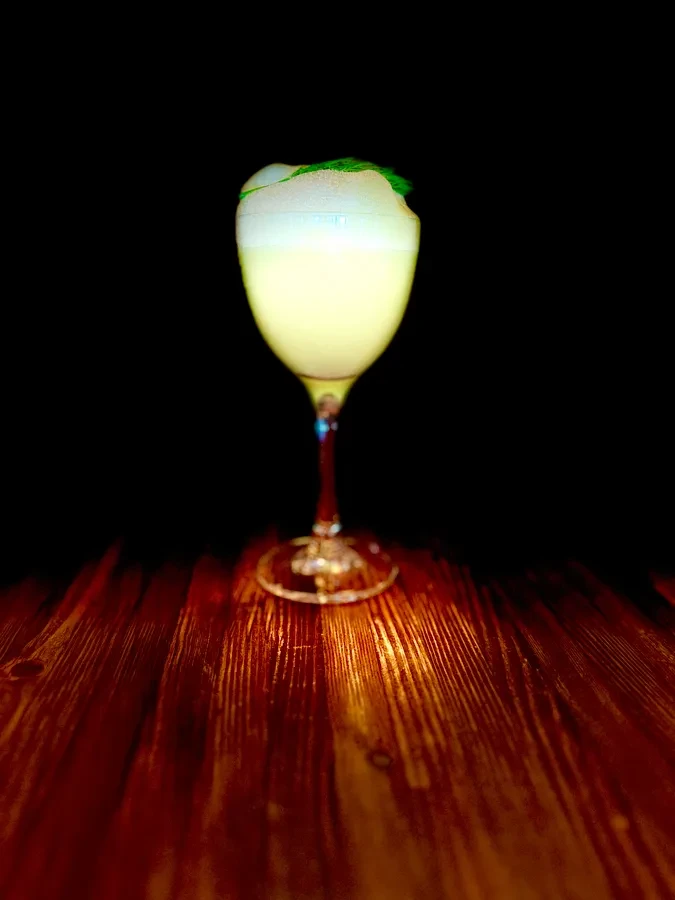 Pale yellow cocktail, foam, green leaves, in stemmed glass on wood.
