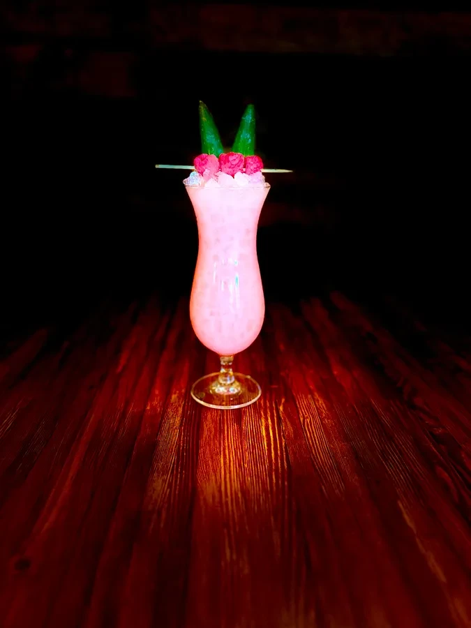 Pink cocktail in hurricane glass with raspberries and green leaves on wood table.