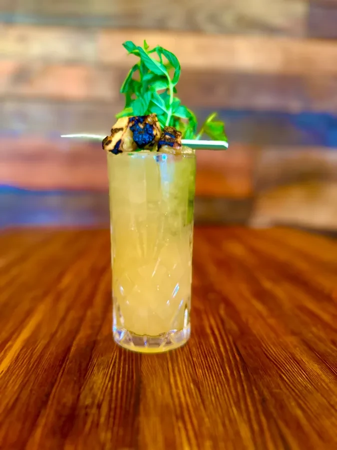 Yellowish-green cocktail with mint and skewered items in a textured glass.