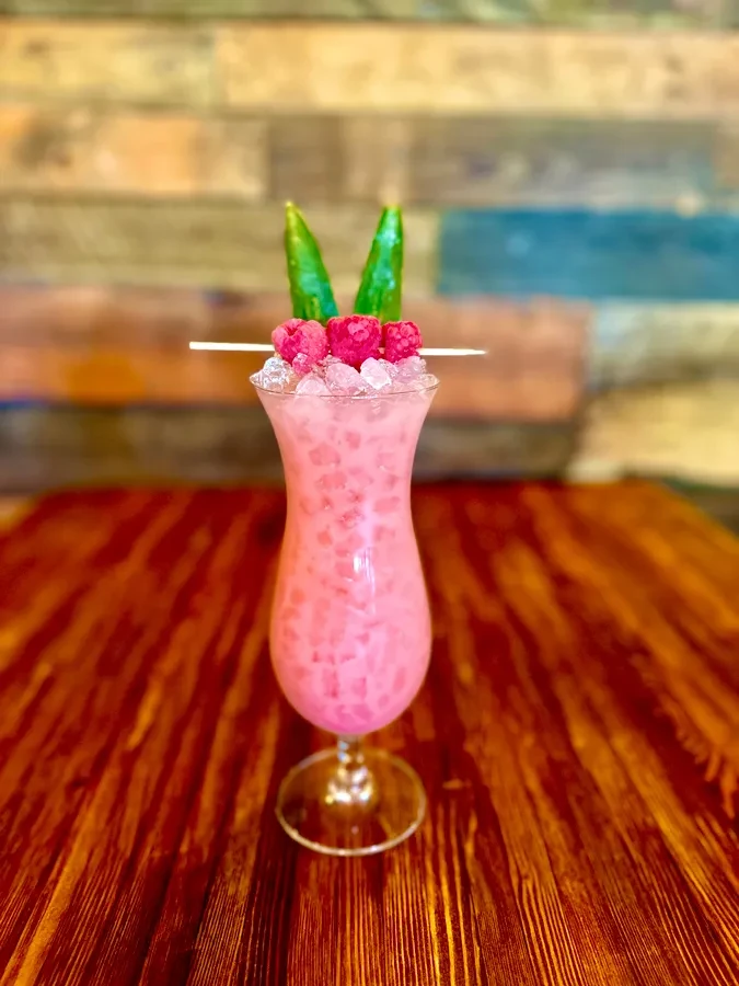 Pink cocktail in hurricane glass with raspberries and green garnish.