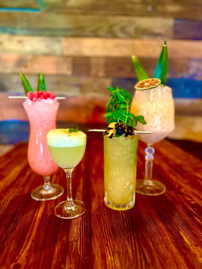 Four distinct, brightly colored cocktails with varied garnishes on a dark wooden table.