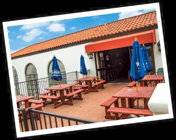 Empty restaurant patio with red picnic tables and blue Modelo umbrellas.
