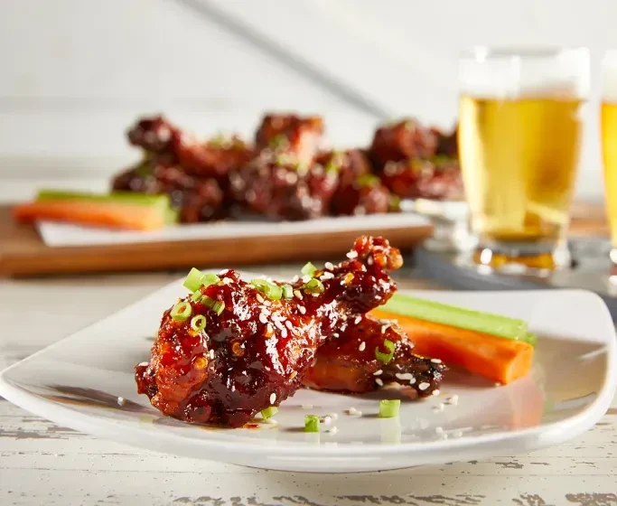 Plate of glazed chicken wings, vegetables, and beer on wood.