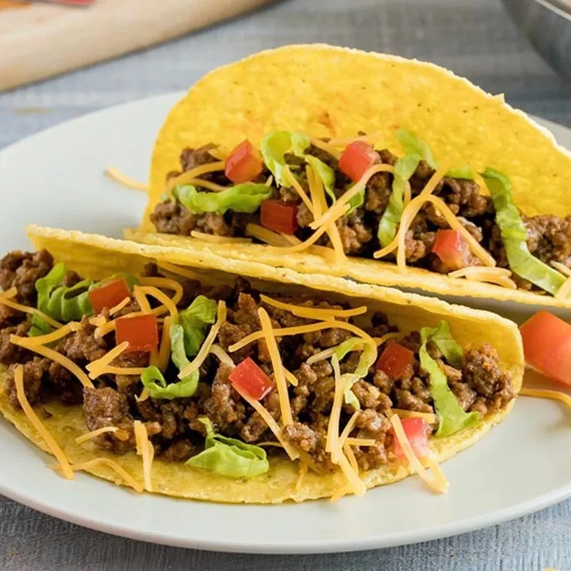 Two hard-shell beef tacos with lettuce, tomatoes, cheese on white plate.