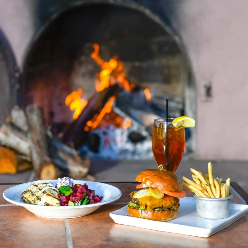 Burger, grilled chicken, fries, and drink by a rustic fireplace.