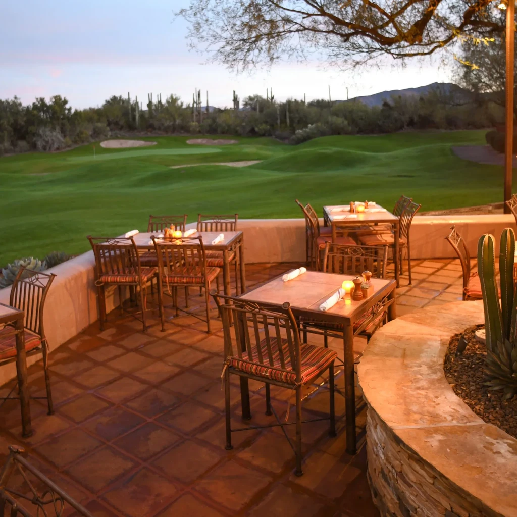 Empty patio dining tables overlook a desert golf course at twilight.