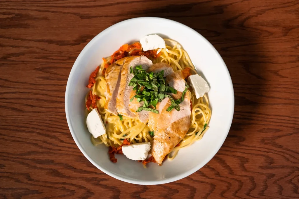 Overhead pasta with chicken, sun-dried tomatoes, herbs, and cheese on wood.