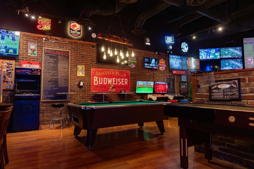 Dimly lit sports bar with a prominent pool table, arcade, and neon signs.