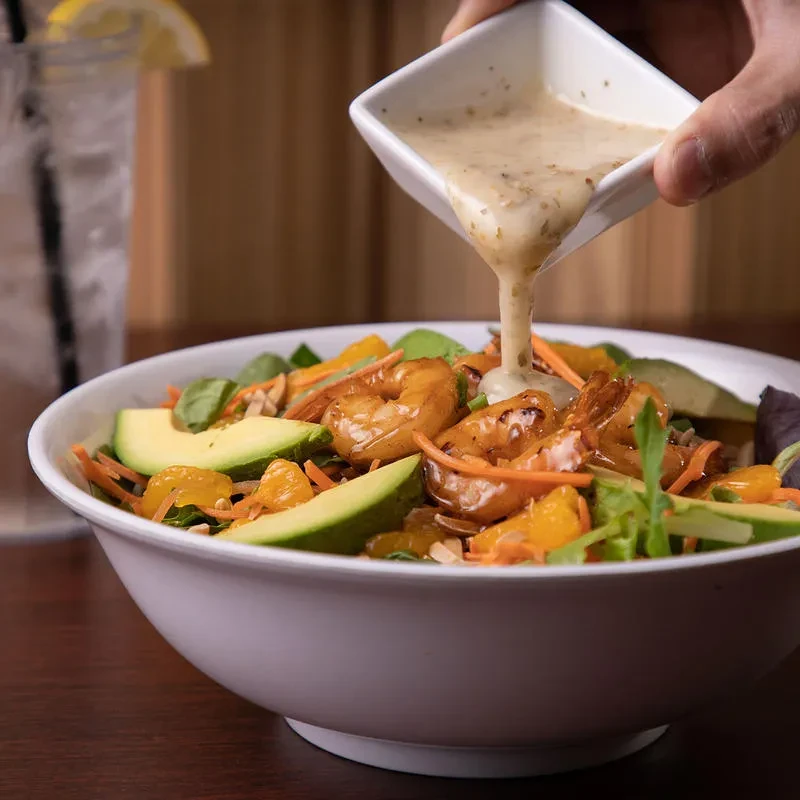 Hand pours dressing on vibrant shrimp, avocado, mandarin salad.