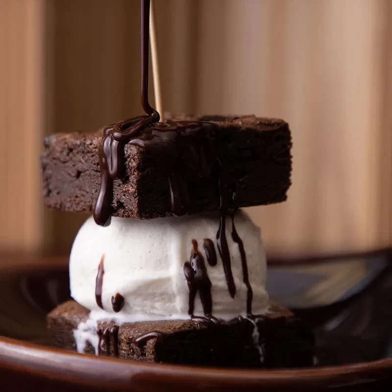 Two fudgy brownies, vanilla ice cream, chocolate sauce being poured.