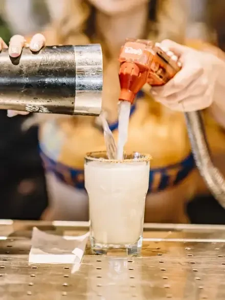 Two hands simultaneously pour a creamy drink into a rimmed glass.