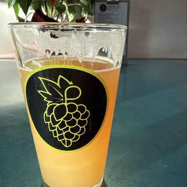 Glass of hazy amber beer, hop logo, foam lacing, teal table.