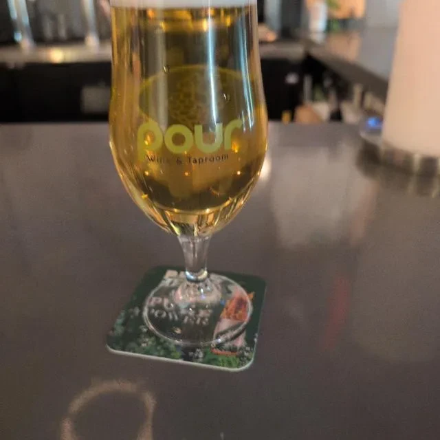 Glass of amber liquid, 'pour Wine & Taproom' logo, in a dim bar.